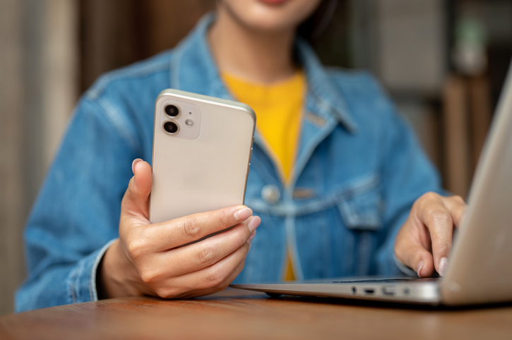 A woman holding her smartphone while working on her laptop, working remotely from a coffee shop.