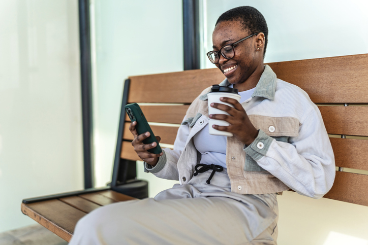 Young stylish black woman with mobile phone, holding coffee waiting for subway train while sitting at train station and texting or watching online video.