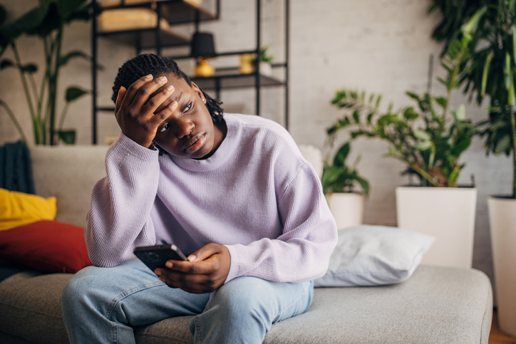 Pensive young woman sitting on sofa with smartphone