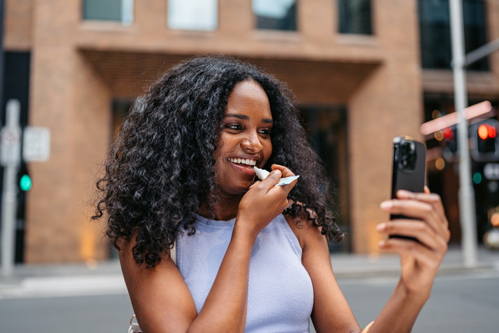 Young Black Woman Applying Lipstick While Looking At Her Reflection On The Phone Screen On The Street In Sydney In Australia